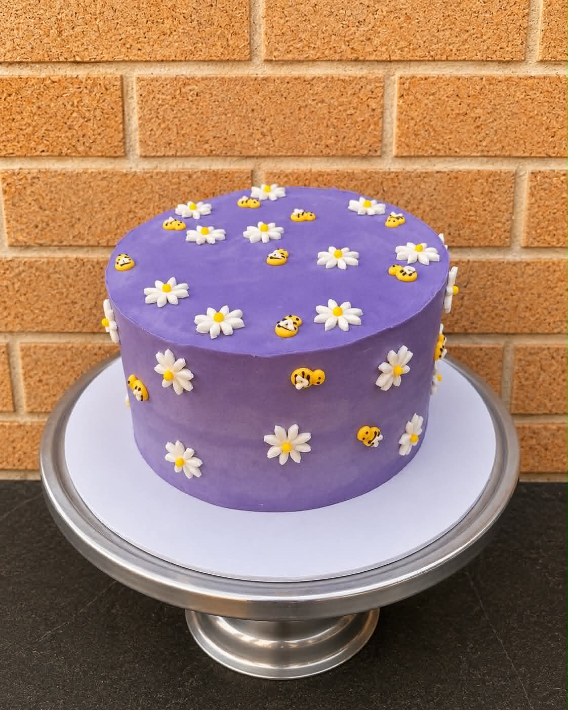 Purple buttercream birthday cake covered all over in white fondant daisies with yellow centres and small yellow fondant bumblebees